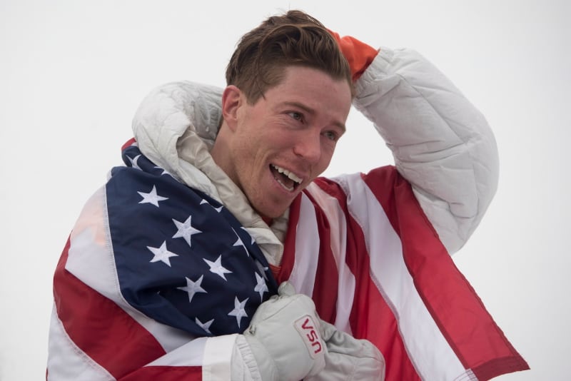 Shaun White smiling in winter gear, standing near a snow-covered halfpipe with mountains behind him