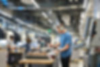 People working in a tech workshop with machines and computers. One person in headphones focuses on a task. Fluorescent lights overhead.