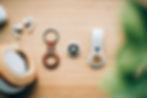 AirPods, a leather keychain, and an AirTag on a wooden surface. Green foliage in the foreground adds a natural touch.