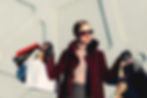 Woman in burgundy coat and sunglasses holds multiple shopping bags. Modern building background. Confident expression.