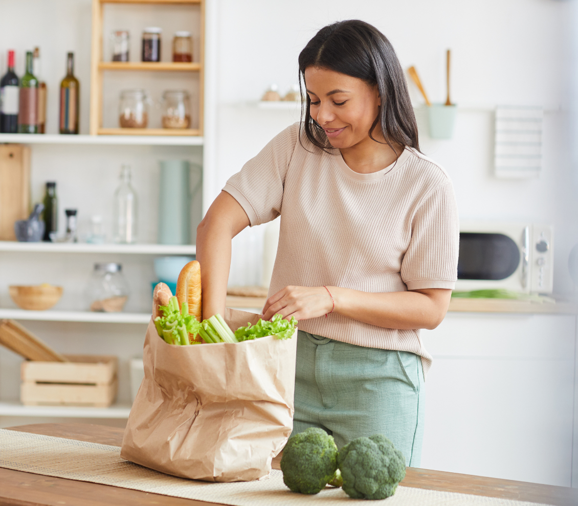 Woman in a bright kitchen sorting fresh produce after grocery shopping, the everyday life that pulls attention away from booking a consultation