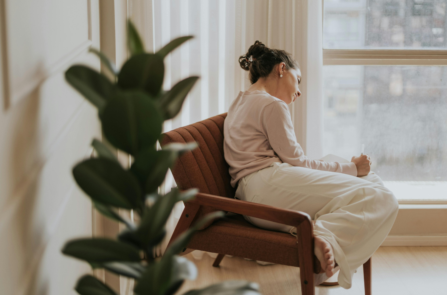 Woman sitting in an armchair by a window, pen in hand, lost in thought while deciding whether to book a consultation