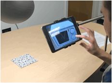 Photo of someone using the AR representation: The marker card is on the table and the participant is viewing the AR representation via a mounted touchscreen.