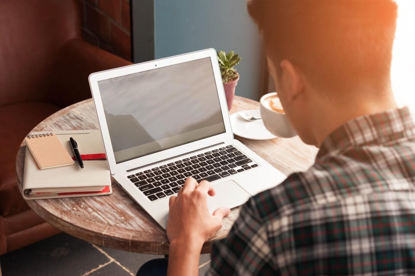 Back view of young man wearing a chequered blue and white shirt, sitting down next to a wooden table and working on his laptop with two notebooks and a pen on his left hand side and holding a cup of coffee with his right hand