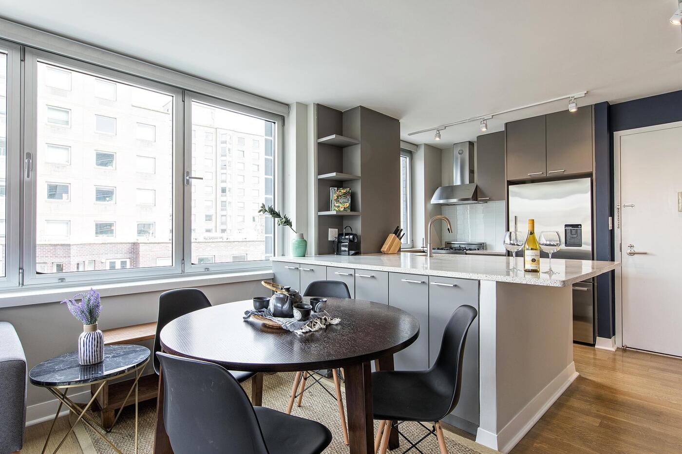 NYC east village kitchen with grey cupboards a dark brown round table with four black chairs a big counter with two glasses of wine and a bottle of wine on it