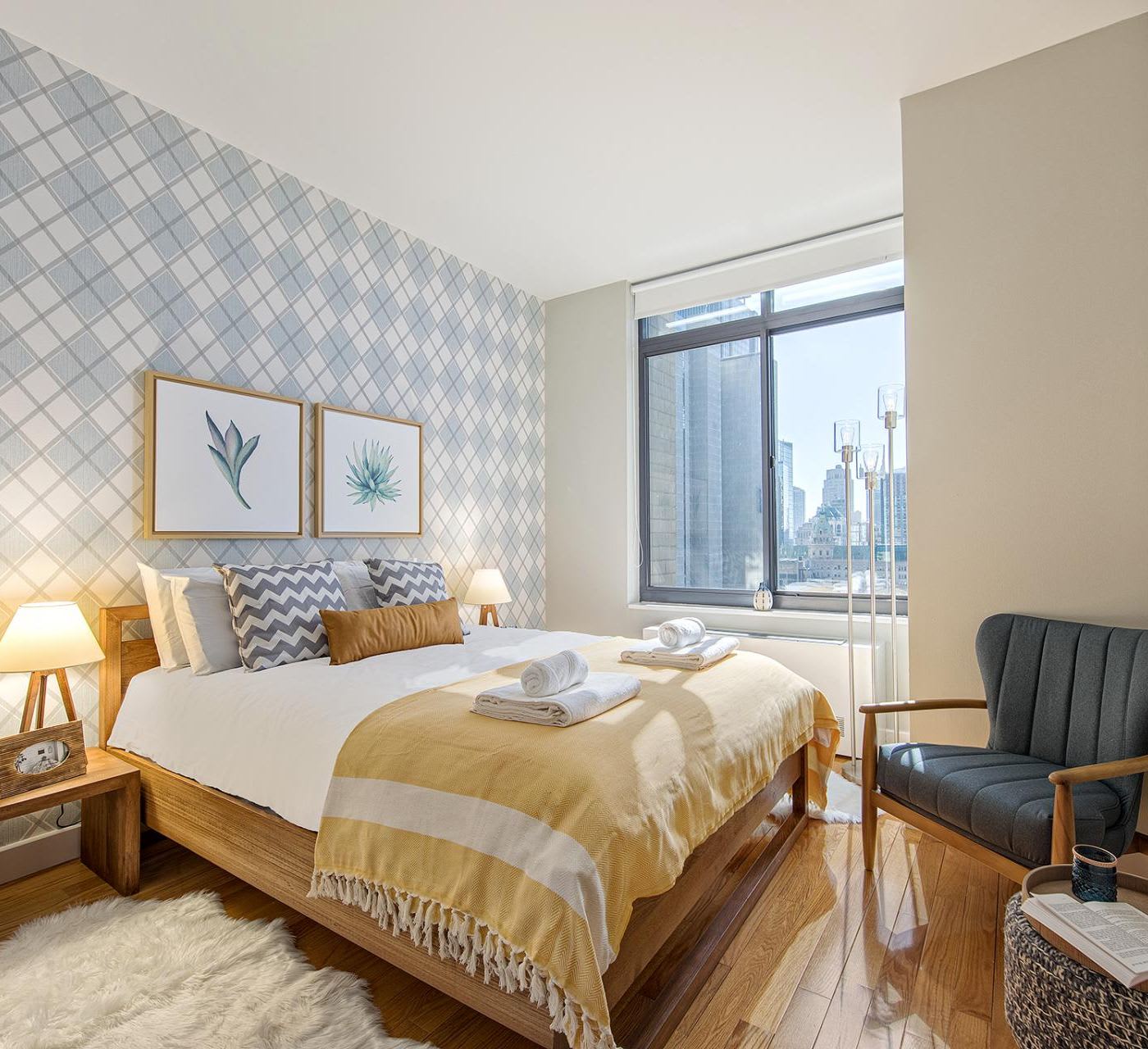 ritz plaza spacious bedroom, teal patterned accent wall above the bed, throw pillows on the bed, wooden bedside table with warm lamp and a dark grey armchair with wooden frame