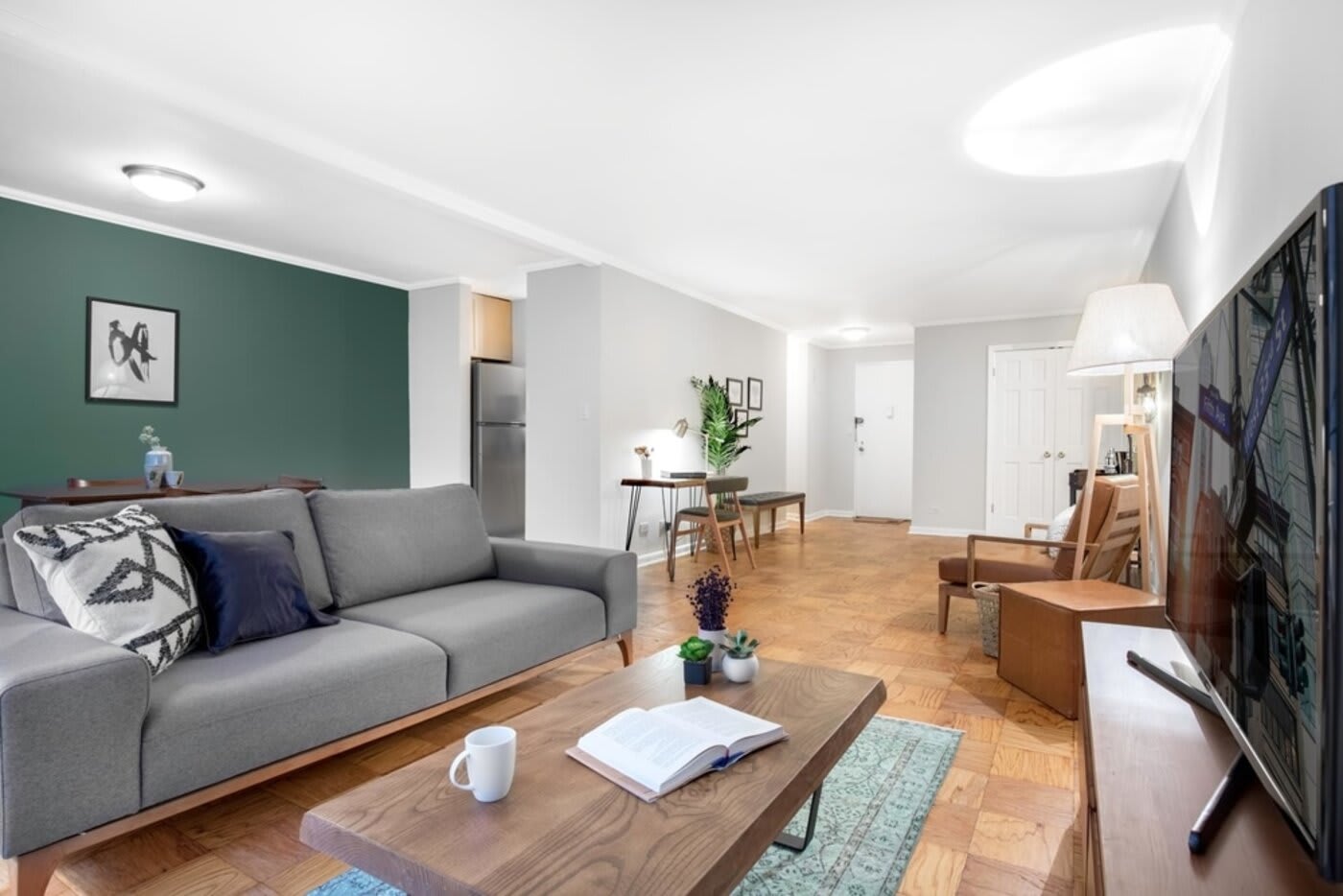 living room with grey couch and wooden coffee table with a white cup of coffee on it a big TV screen white walls one green wall