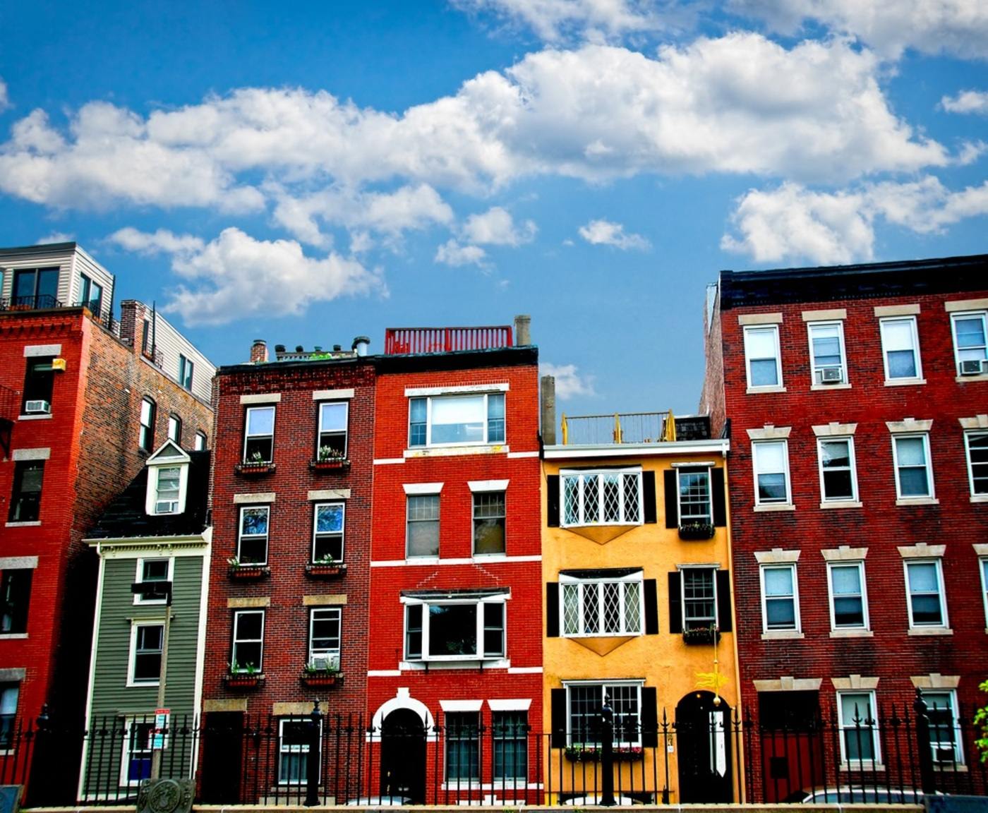 Various brightly colored brick buildings are all next to each other with a different style of windows and flower boxes with a black iron fence in the foreground in front of all of them. There is a green building and a yellow one and the rest are red. Above the buildings is a blue sky with white clouds.