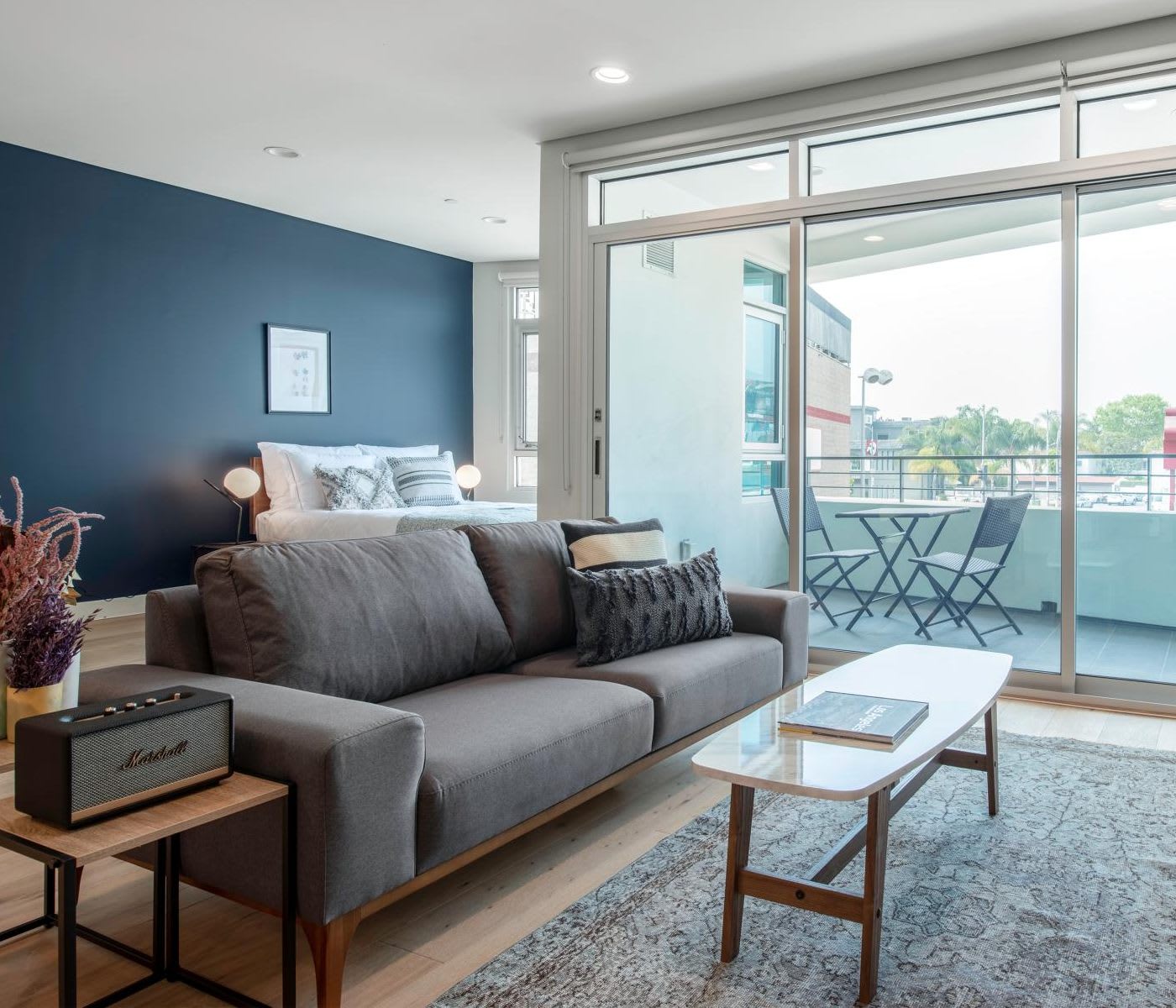 studio apartment with a grey free-standing couch, a side table with a marshal speaker on it, a bed further back and a balcony with a small table
