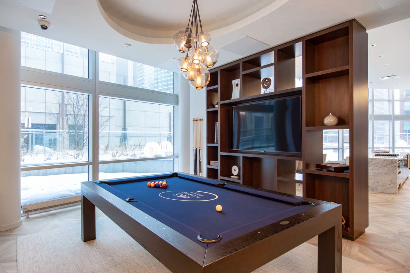 common area in an apartment building with a wooden pool table in front of large panel windows and a dark wooden shelves unit
