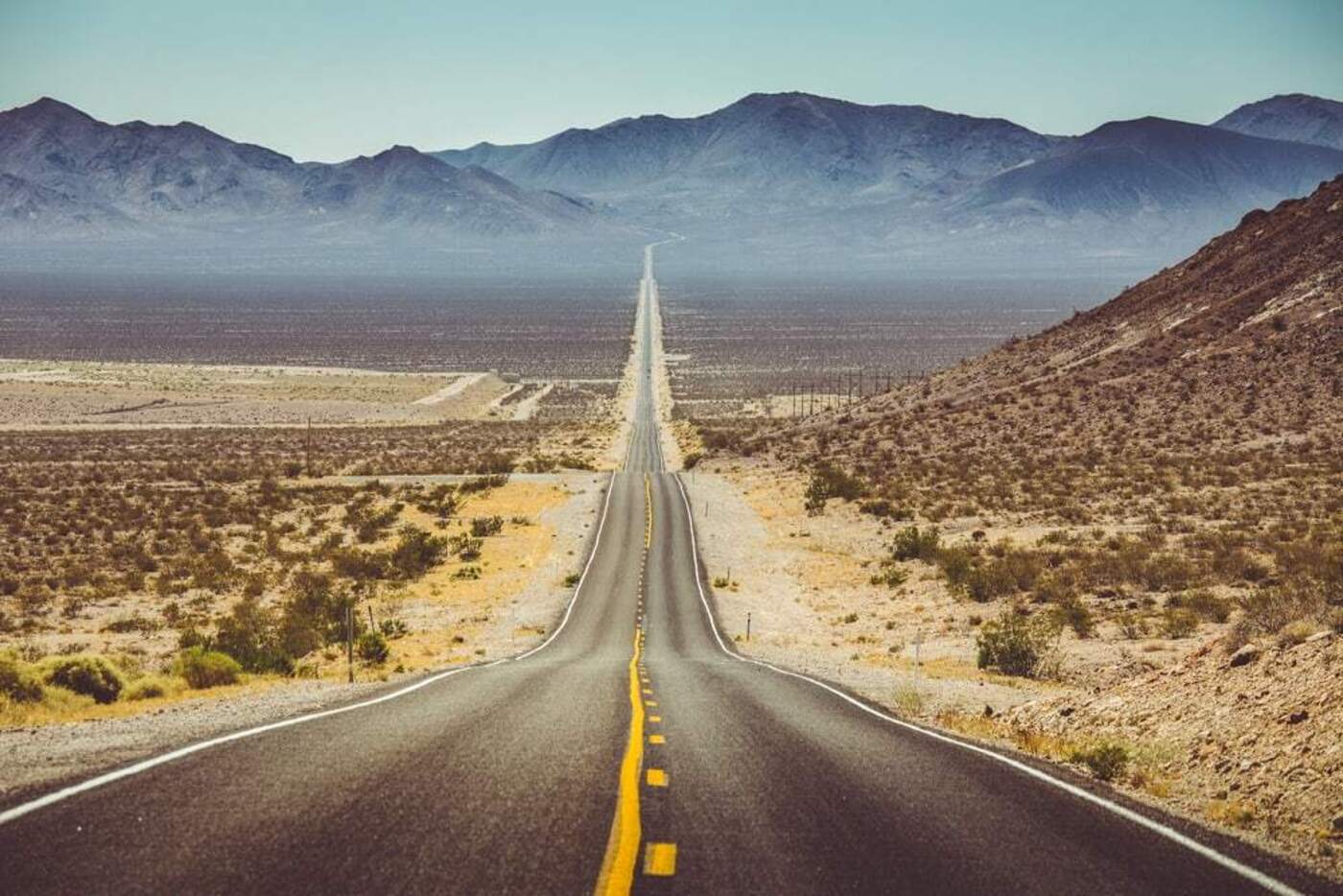 wide open black red with yellow lines down in the middle going through the desert with mountains in the background and brush on either side of it on the West Coast of the USA