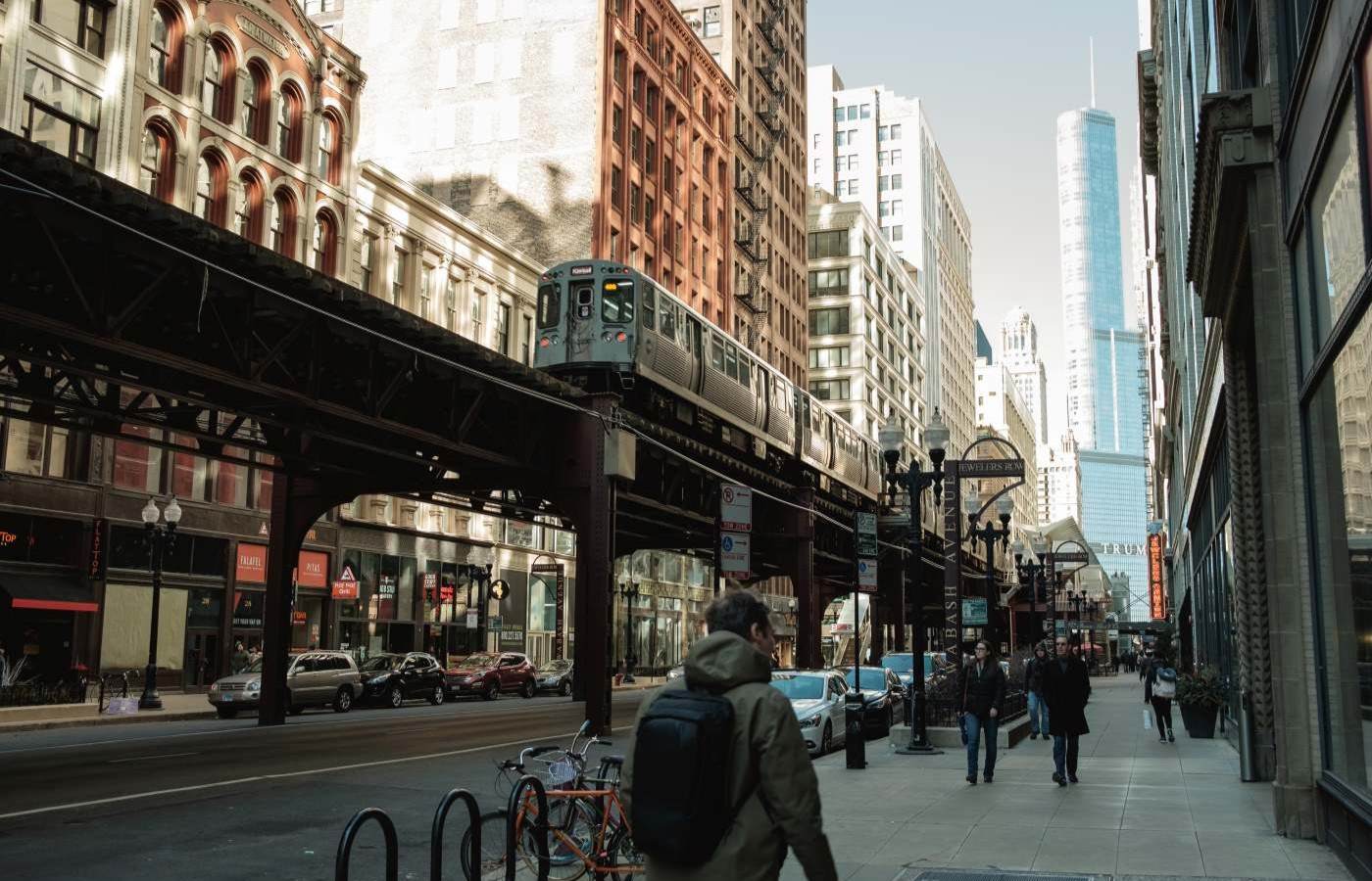 A man walking on a sidewalk of a chicago street. A train can be seen on a raised platform above the road and tenements line both sides of the street 