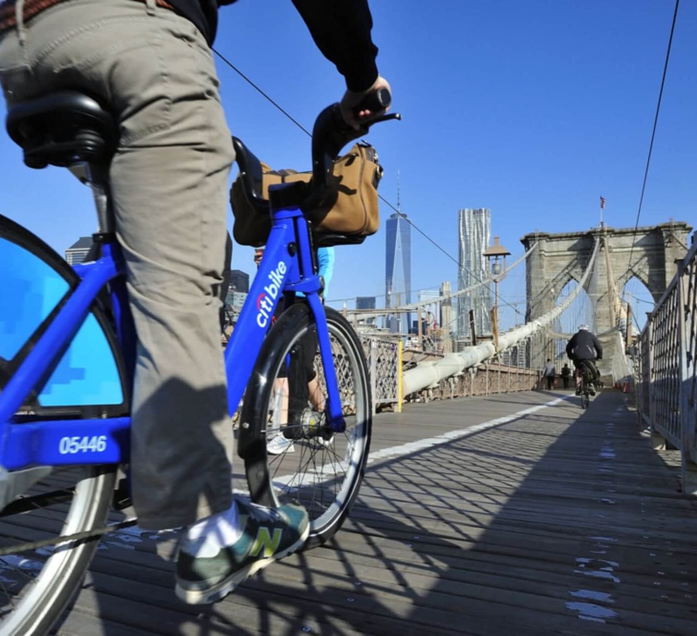 man with grey pants and black shirt rides a blue citi bike down the brooklyn bridge