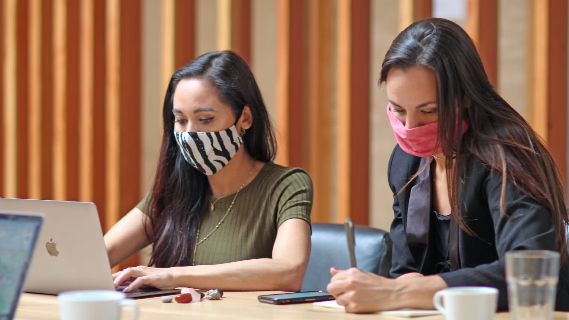 To women wearing medical masks working side by side in a coworking space.