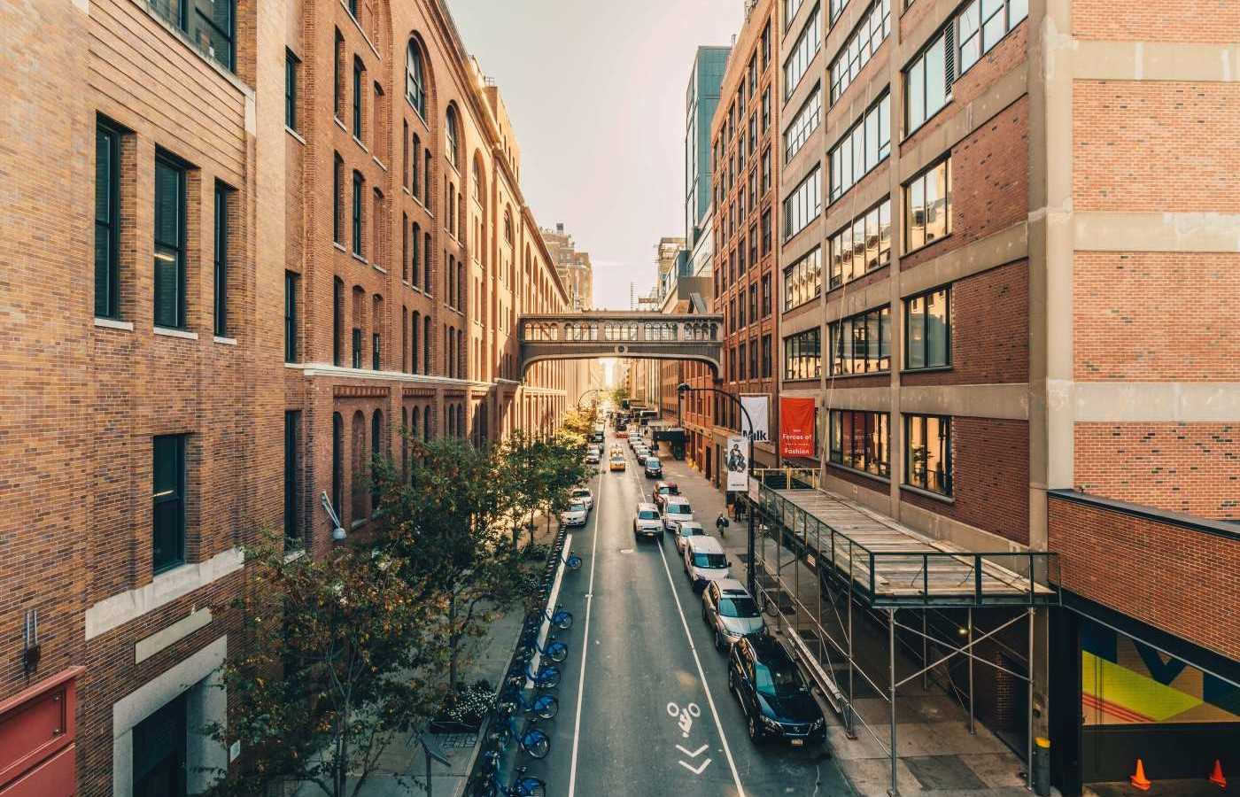 A new york street flanked by multi-story brick buildings. A pedestrian bridge can be seen in the distance.