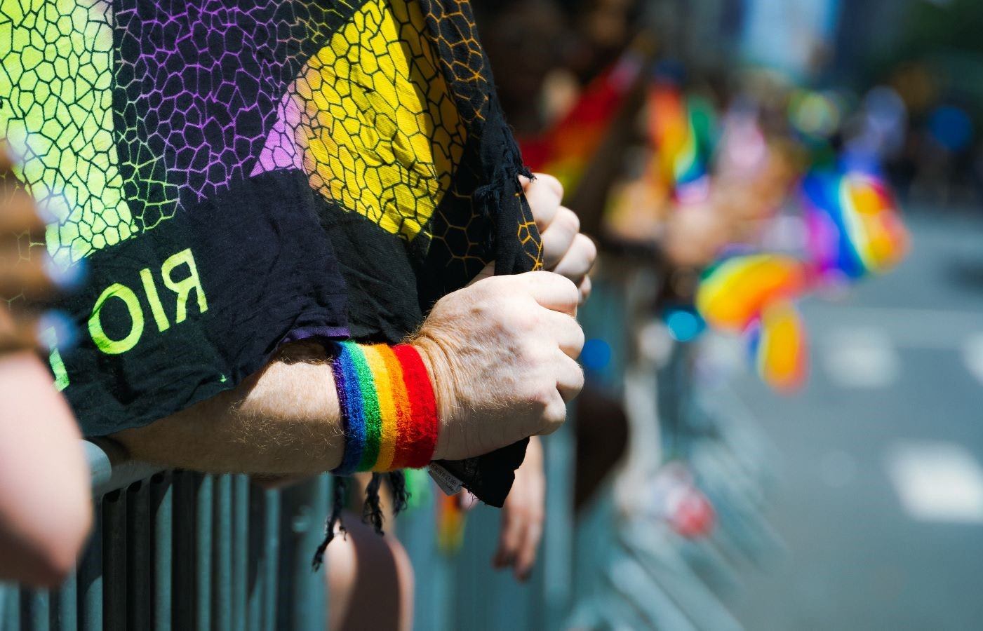 People leaning onto a metal street barricade holding pride flags during a gay pride event.