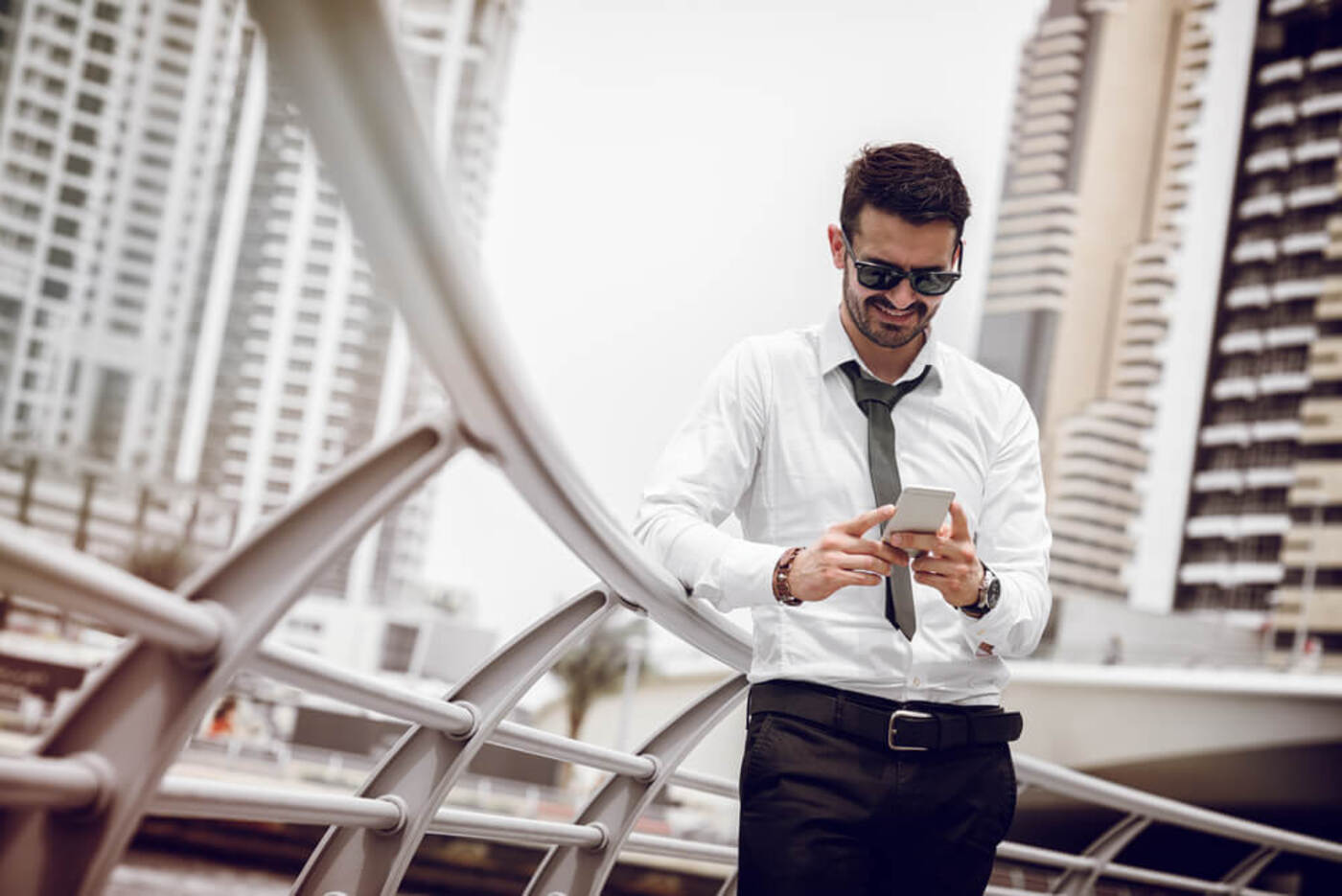A man wearing a white shirt and a black tie and black pants and dark sunglasses holds and looks at his mobile phone while leaning against a railing in Dubai