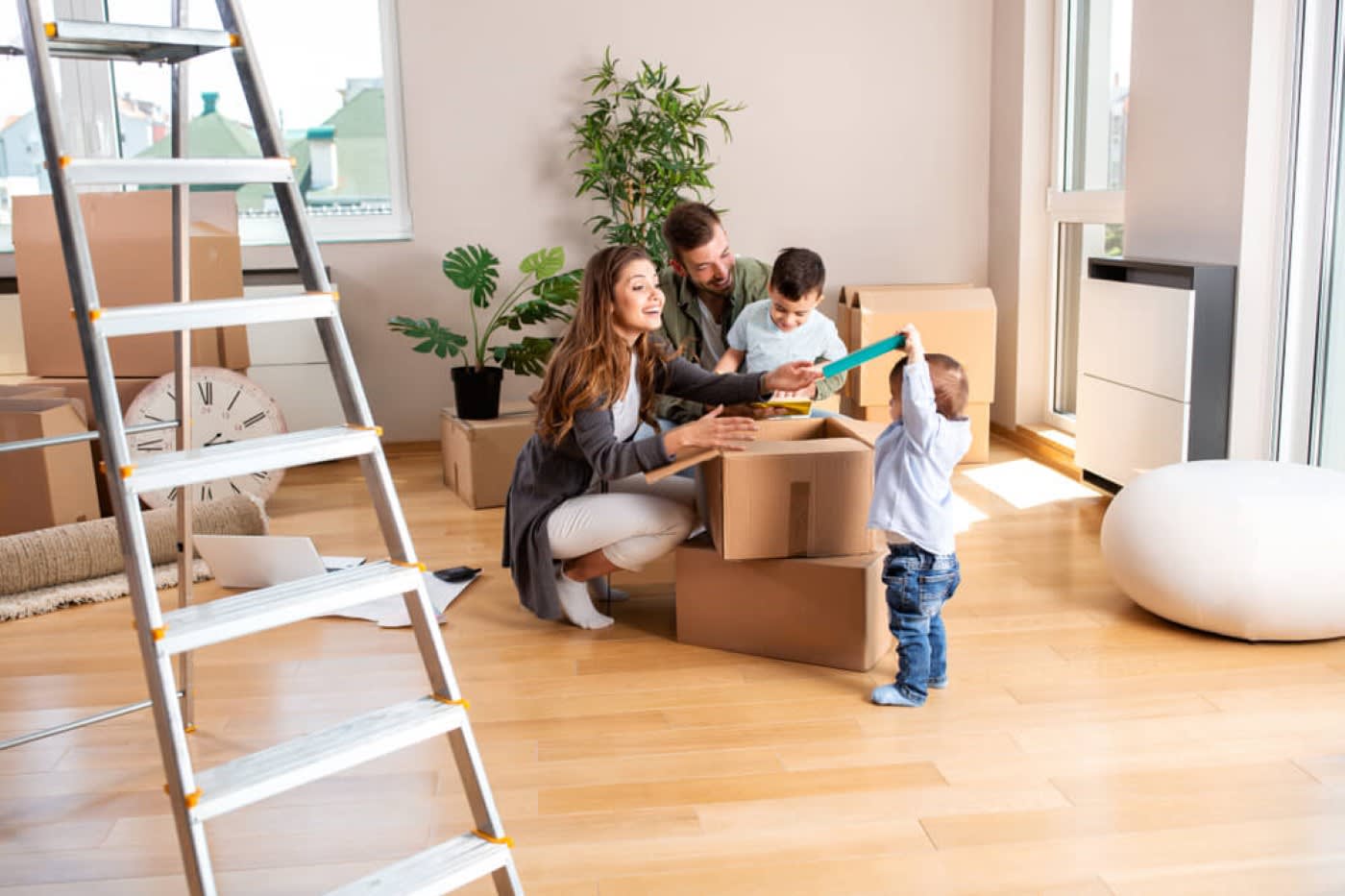 A mother and a father and two young sons are sitting in the middle of the room around packing boxes after moving into a new home. They are in the middle of unpacking the boxes together. There are some plants behind them as well as more boxes. In the foreground of the photo is a tall ladder.
