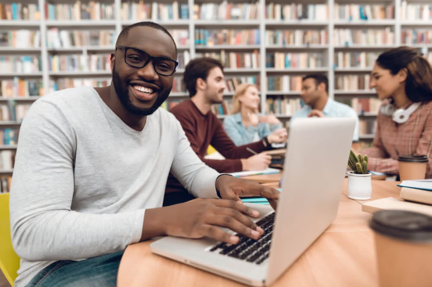 A student is wearing a gray shirt and smiling while working on his silver laptop. Behind him there are other students talking and behind them there is tall bookshelf going across the entire wall.