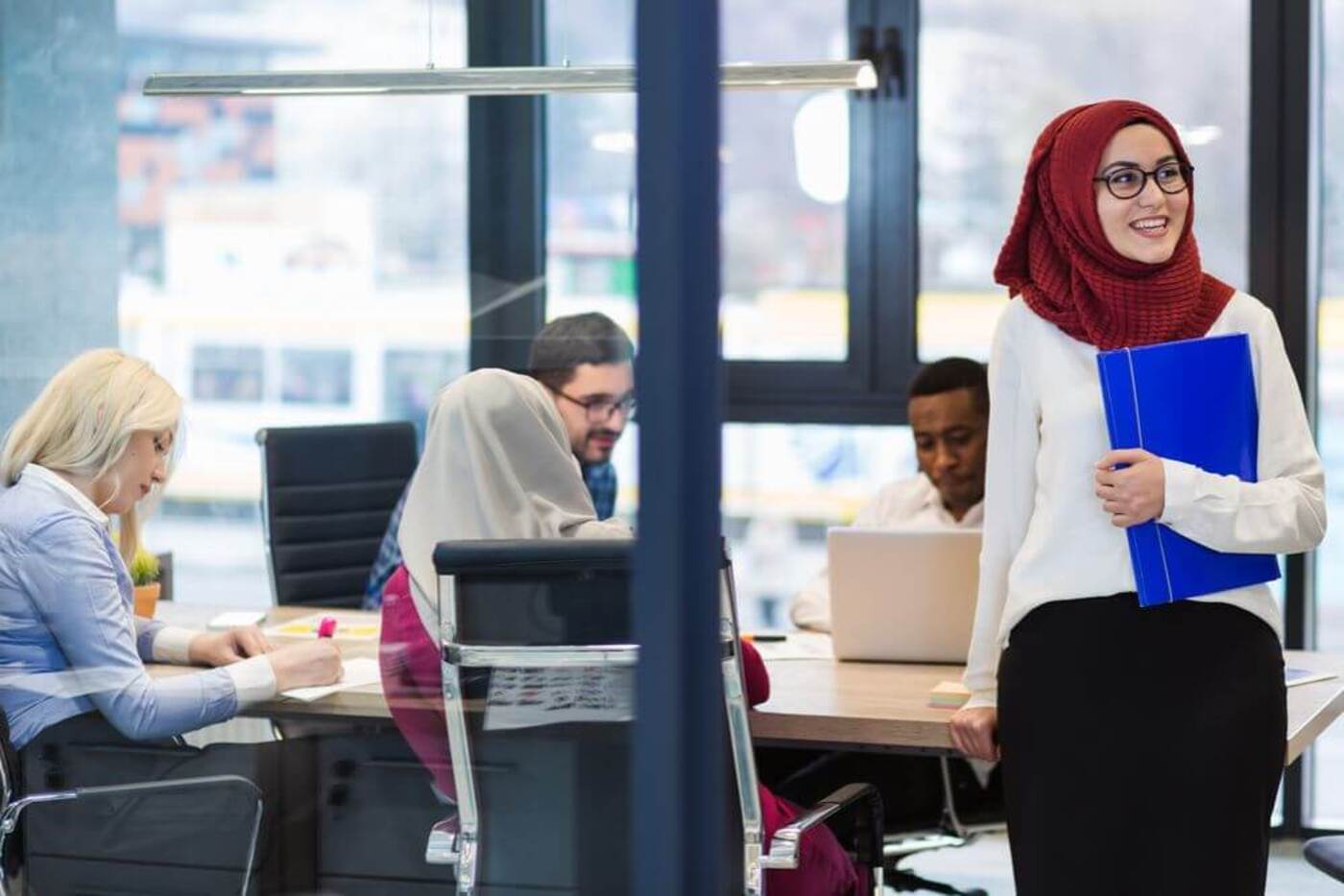 A woman wearing a red veil and a white long sleeved shirt is holding a blue folder. Behind her is a table of men and women having a meeting