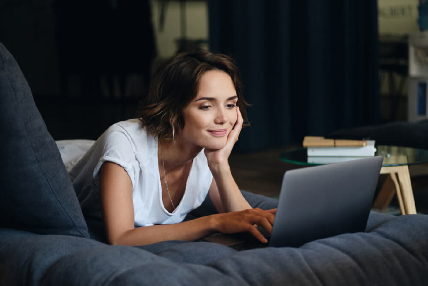 A girl with brown hair wearing a white shirt is laying on a gray couch working on a silver computer. There is a small round table next on her left side