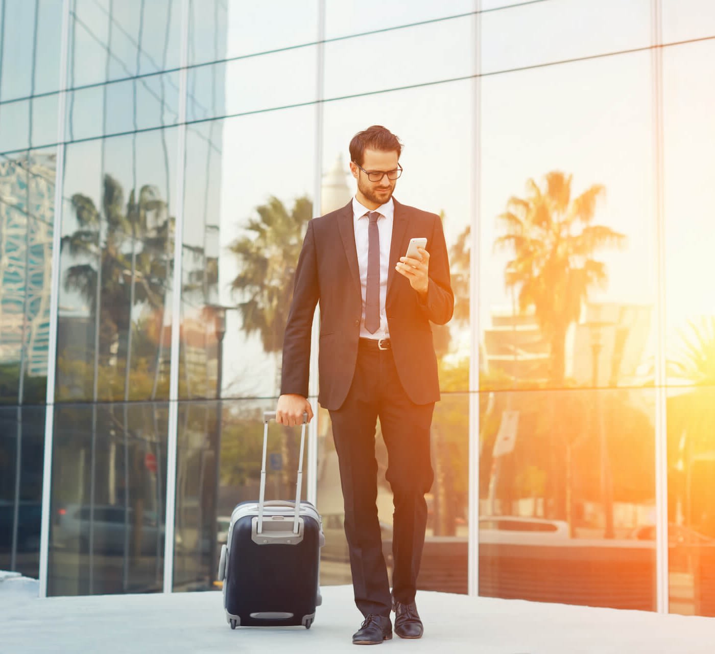 man dressed professionally looking at his phone while carrying a carry-on suitcase at the airport