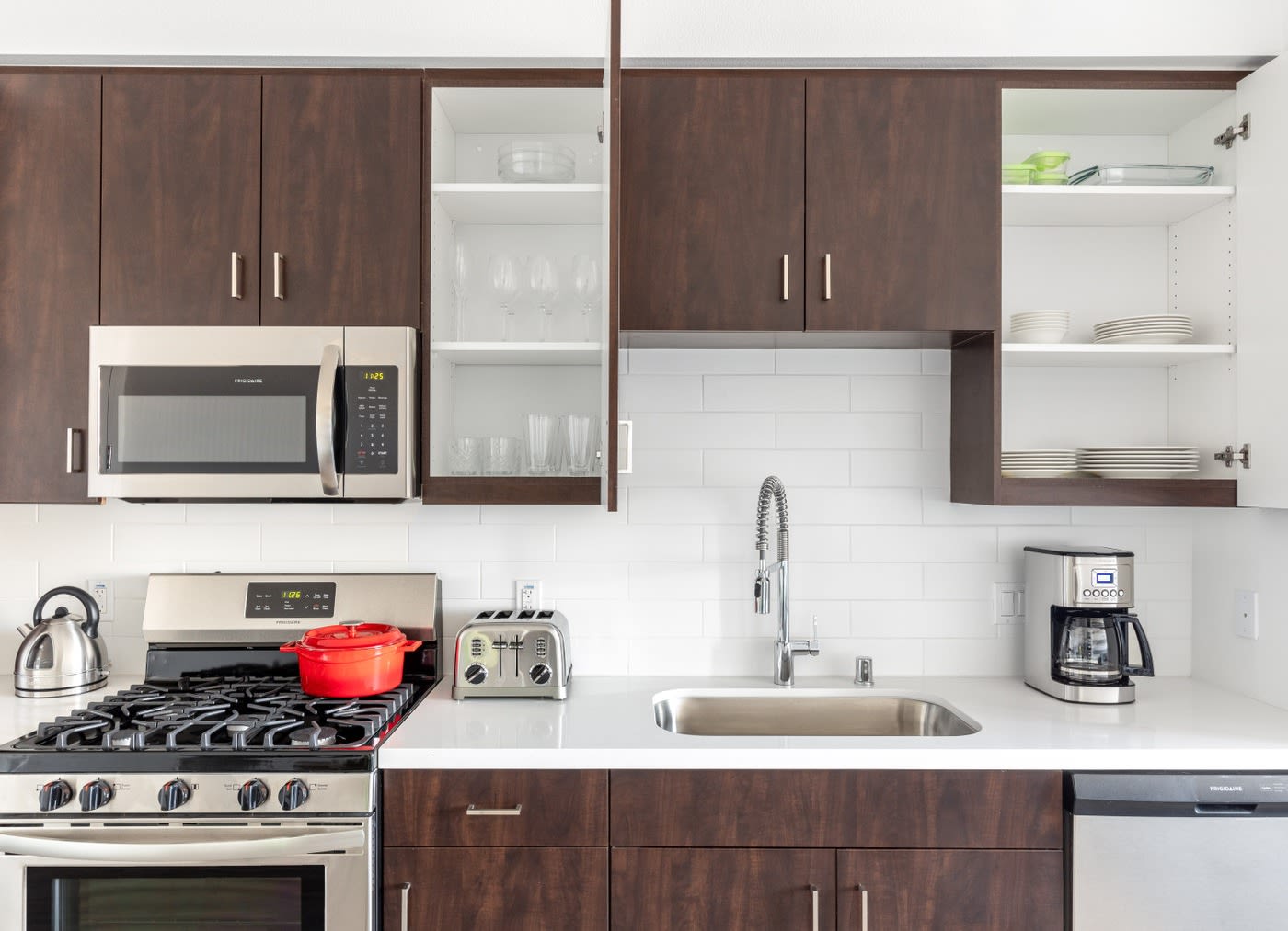 A fully equipped kitchen with dark wood cabinets and plates and cups on each shelf. There is a red pot on the stove and a white coffee maker on the counter
