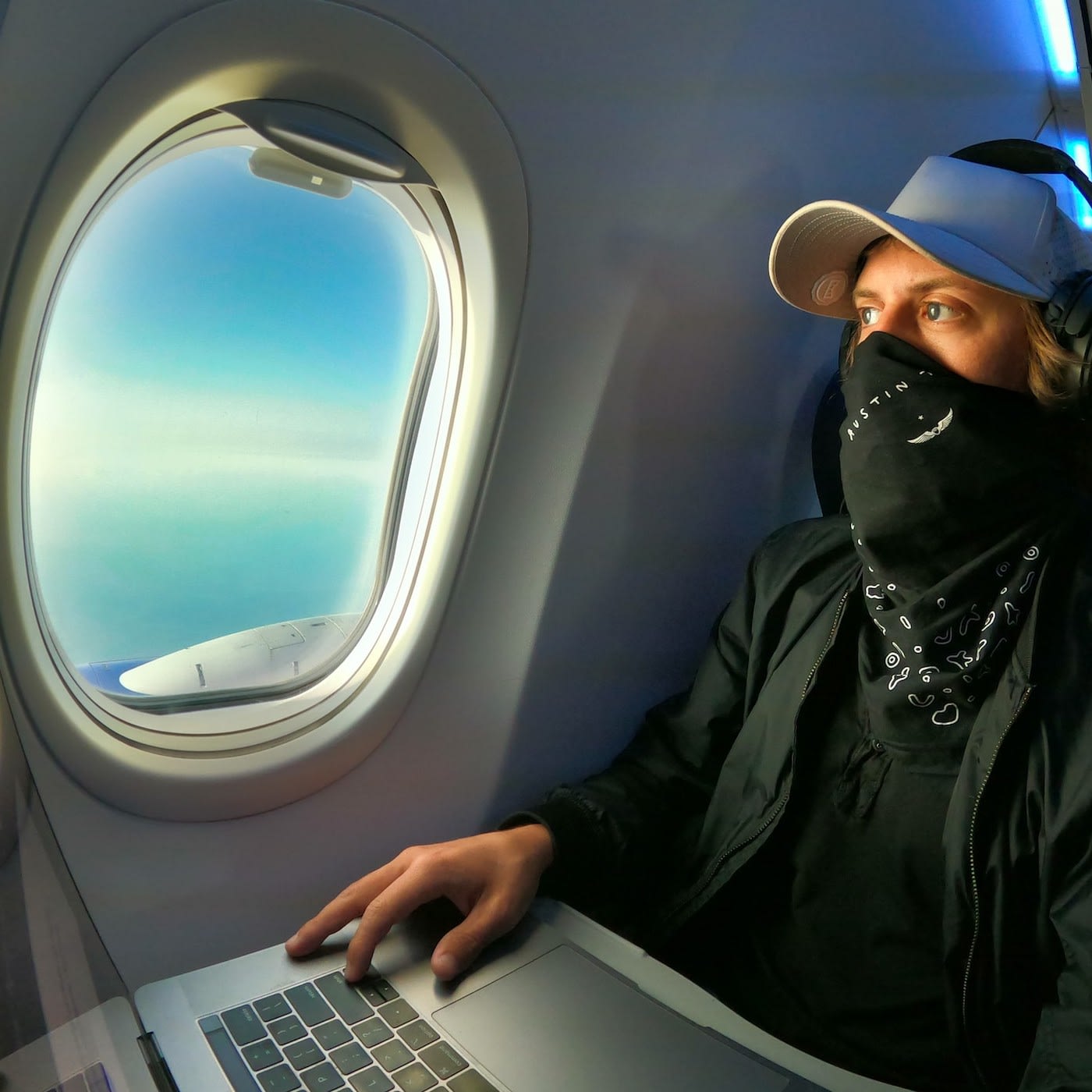 man wearing a mask looking out the airplane window with a laptop in front of him