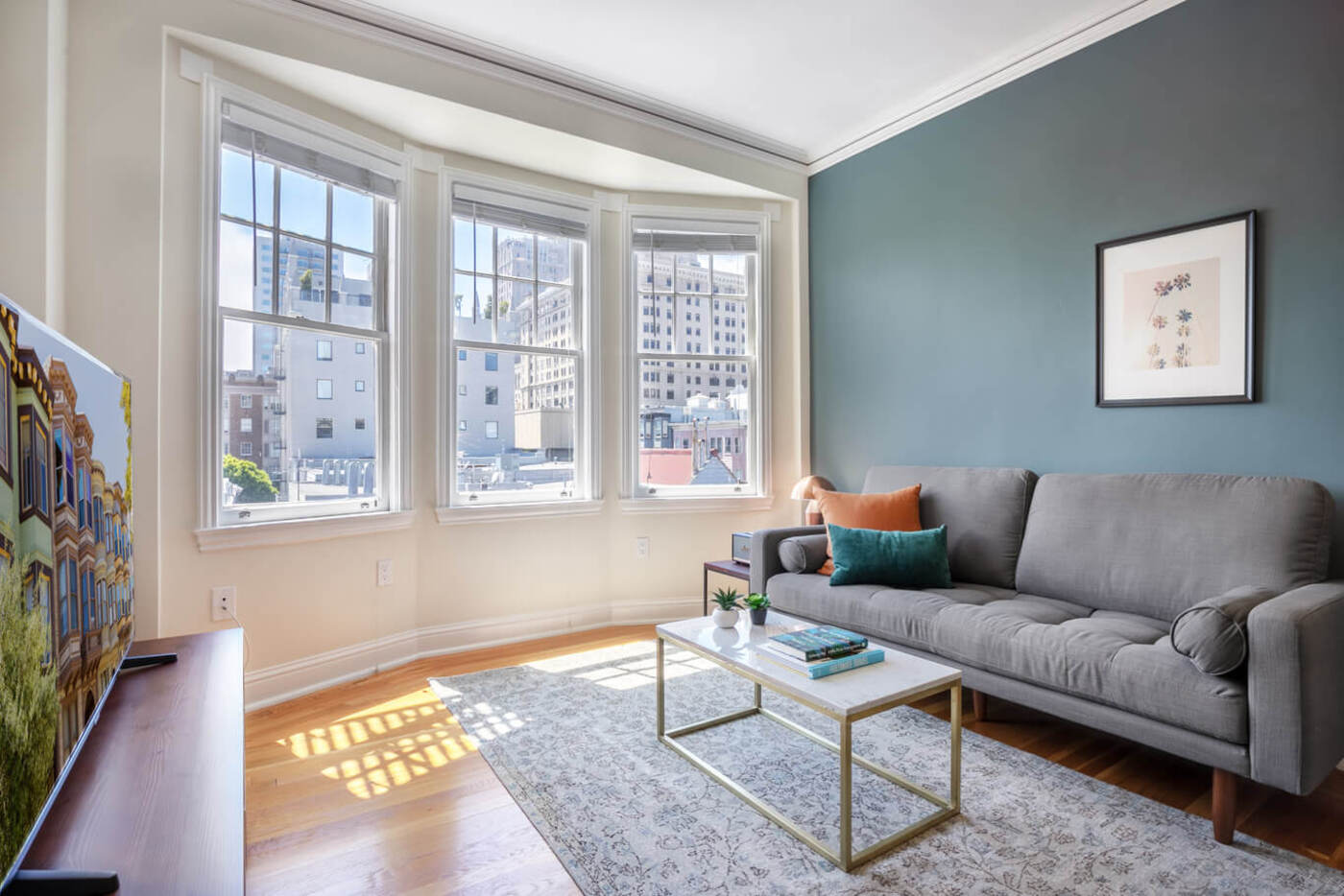 minimal apartment with three large windows letting natural light in, green accent wall behind the grey couch and a small marble coffee table