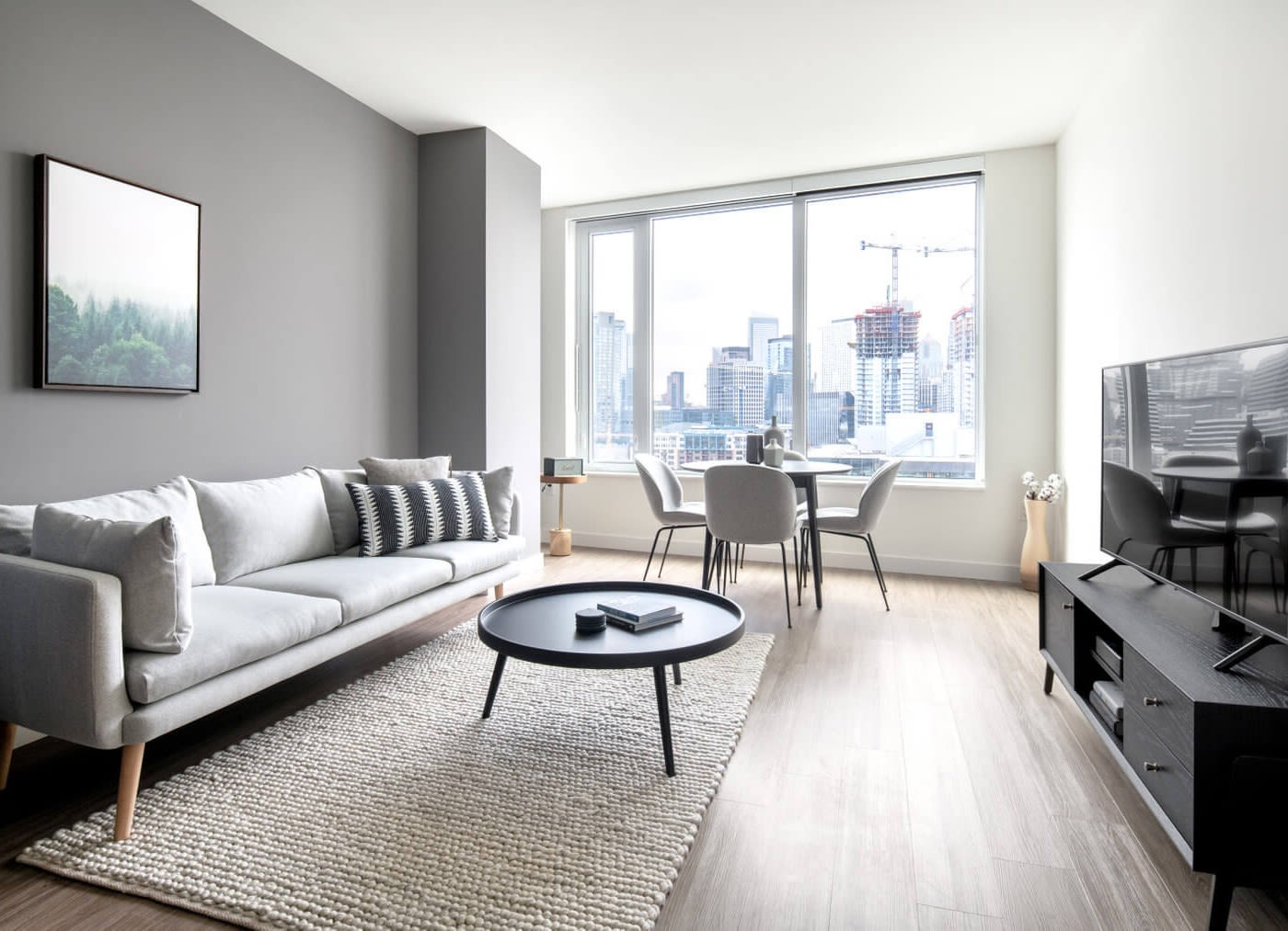 Inside view of an apartment with wooden floor and a beige carpet on the left half of the floor. On top of the carpet is a a black coffee table and a grey sofa with a blue and white striped cushion and behind the couch is a grey wall with a small photo frame of the woods which is opposite a black flat screen television on top of a black tv stand. In the background is a table with four chairs next to a large window with a view of the city