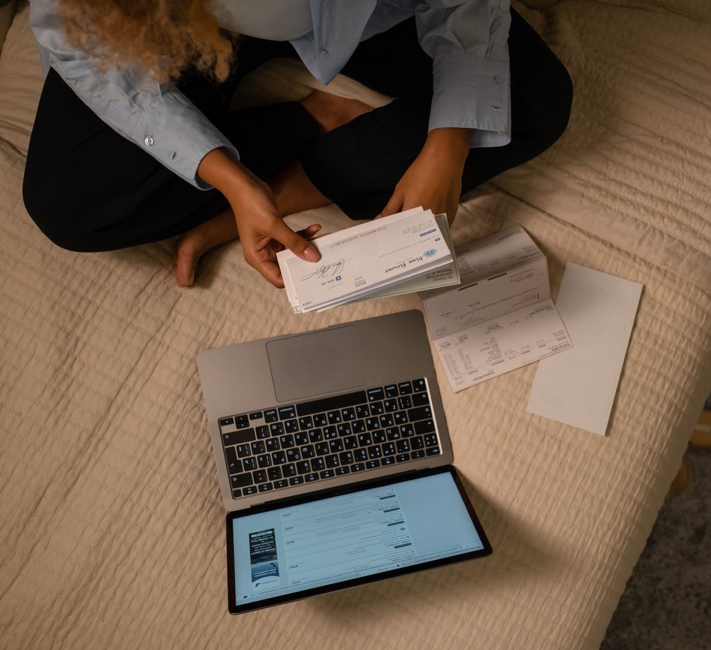 woman sitting on a bed with her laptop and other papers, holding a cheque