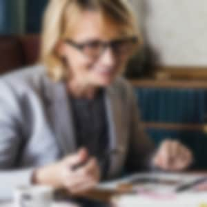 A woman with a grey suit jacket and black glasses is holding a pen and smiling as she sits across from a woman with black hair wearing a black shirt. They are looking at some papers on the table in front of them