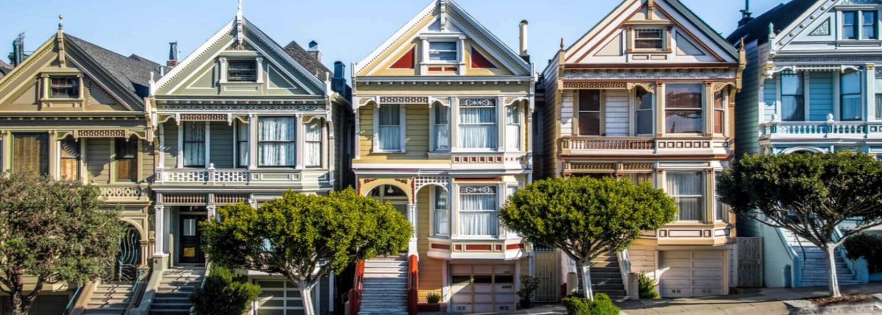 The multicolored houses in a row in San Francisco that is called the Painted Ladies. The street goes up at an angle and there are trees in front of the homes with a blue sky behind them