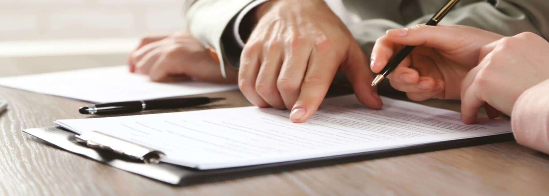 a man sitting a wooden table pointing at a piece of paper on a clipboard.