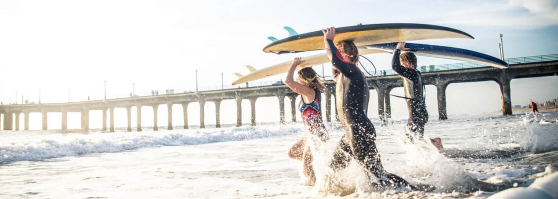 three people holding surfboards above their heads walk out into the water near the pier in Los Angeles