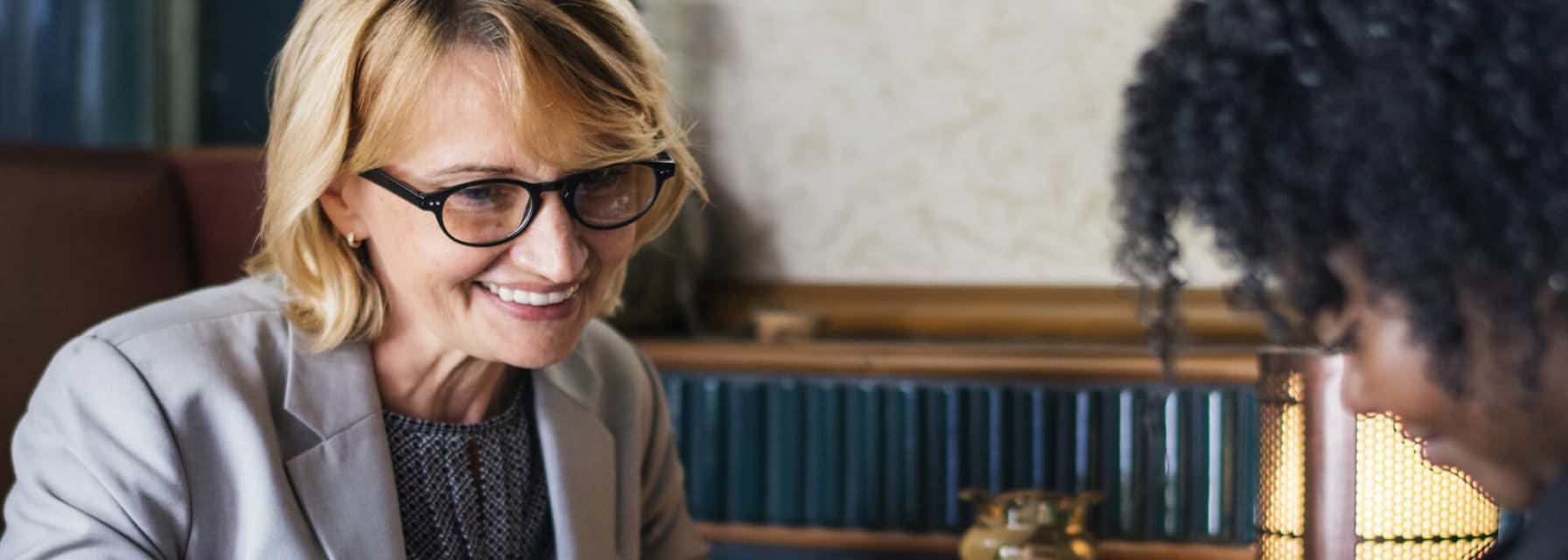 A woman with a grey suit jacket and black glasses is holding a pen and smiling as she sits across from a woman with black hair wearing a black shirt. They are looking at some papers on the table in front of them