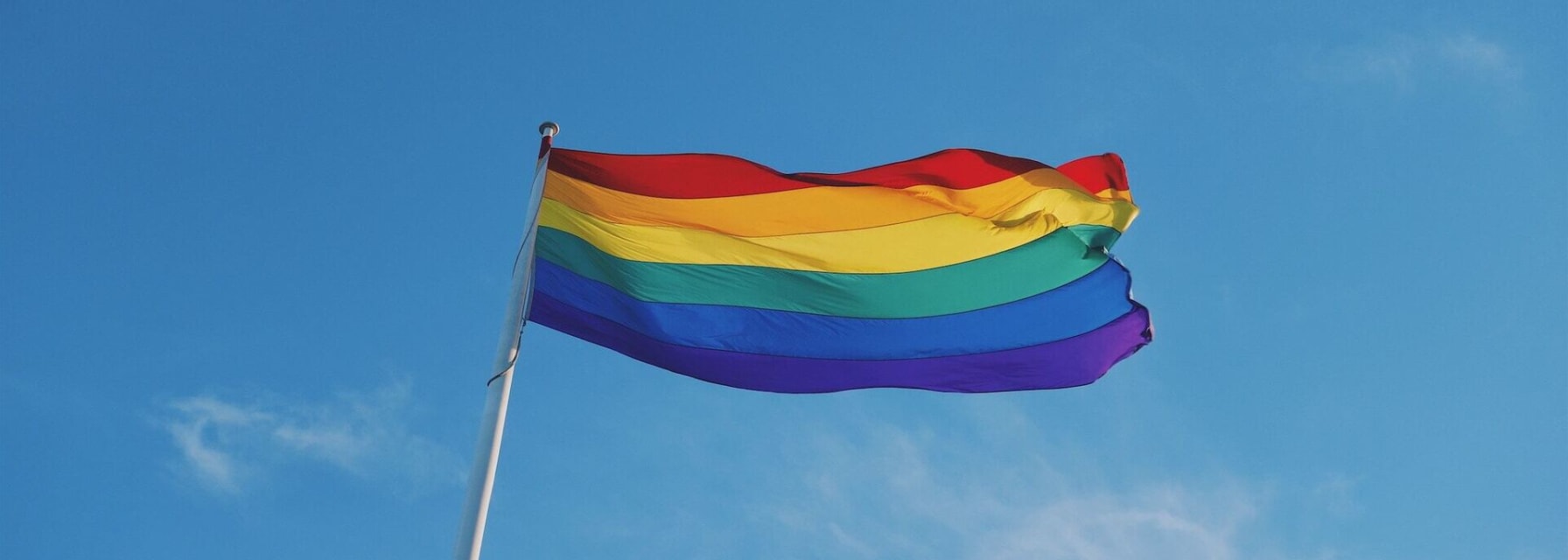 A pride flag waving atop a flagpole as seen from the ground.