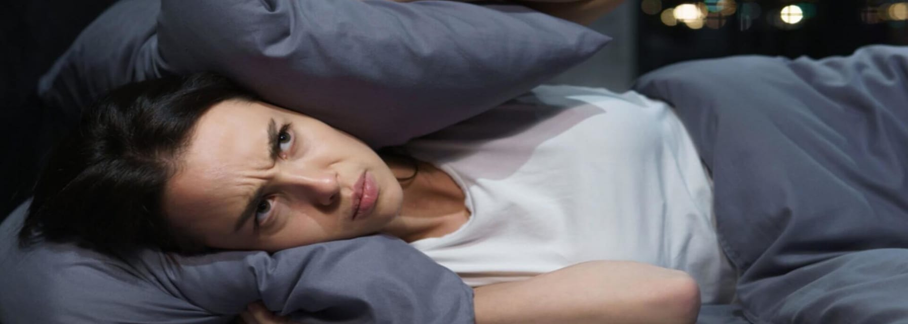 woman laying on her bed looking angry and covering her eyes with a pillow to block out the loud noise