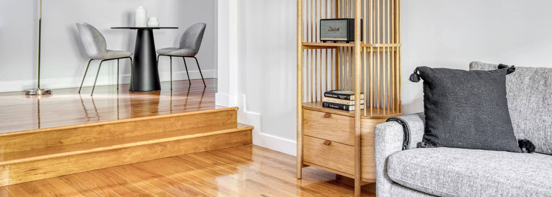 minimal living space, wooden floor, grey couch, wooden shelving unit with very little decor and a small table for two with a muted painting above and a floor lamp next to it