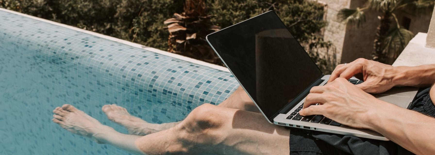 man working with a laptop on his lap sitting on the edge of a pool dipping his feet in