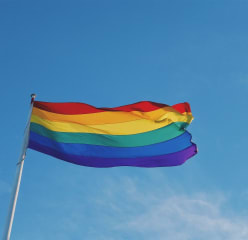 A pride flag waving atop a flagpole as seen from the ground.
