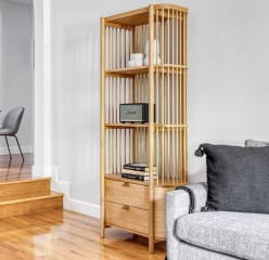 minimal living space, wooden floor, grey couch, wooden shelving unit with very little decor and a small table for two with a muted painting above and a floor lamp next to it