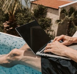 man working with a laptop on his lap sitting on the edge of a pool dipping his feet in