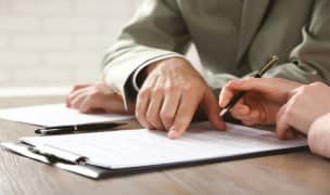 a man sitting a wooden table pointing at a piece of paper on a clipboard.