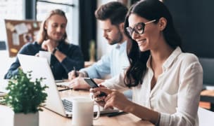 woman sits at her computer while looking at her phone sitting at a table with other people are in a coworking space