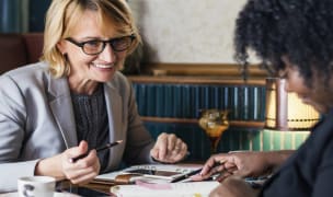 A woman with a grey suit jacket and black glasses is holding a pen and smiling as she sits across from a woman with black hair wearing a black shirt. They are looking at some papers on the table in front of them