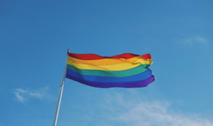 A pride flag waving atop a flagpole as seen from the ground.