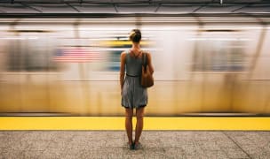 Woman in a grey dress with bare legs standing with her back to the camera, facing a fast moving train