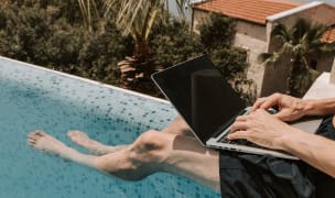 man working with a laptop on his lap sitting on the edge of a pool dipping his feet in