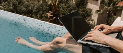 man working with a laptop on his lap sitting on the edge of a pool dipping his feet in